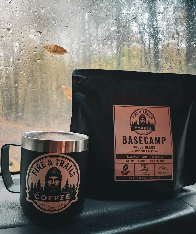 Basecamp coffee bag and a Fire & Trails mug on a vehicle dashboard beside a rain-covered windshield