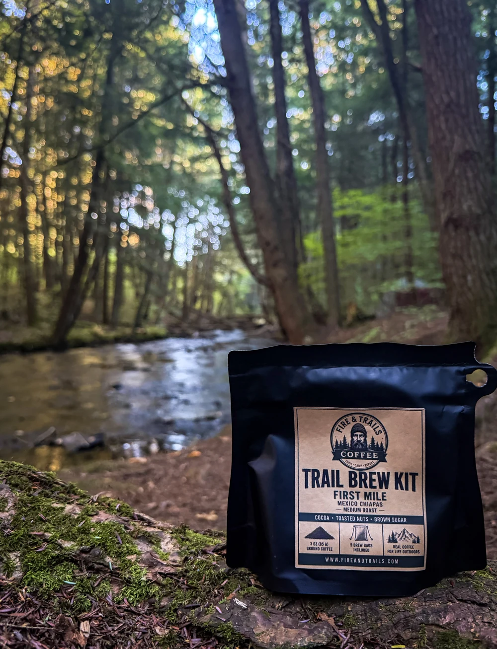 Fire & Trails Trail Brew Kit beside a forest creek with its First Mile coffee label visible