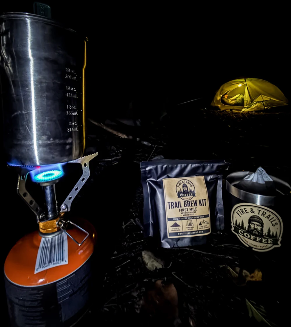Camp stove, Trail Brew Kit, and Fire & Trails mug beside an illuminated tent at night